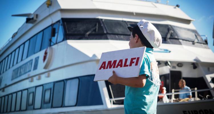 Enfant tenant un panneau "Amalfi" devant un ferry.