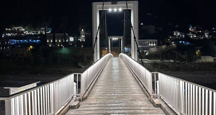 Puente peatonal iluminado de noche con vista de edificios cercanos.