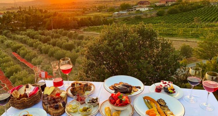 Configuración de comedor al aire libre con comida y vino con vista a un viñedo al atardecer.