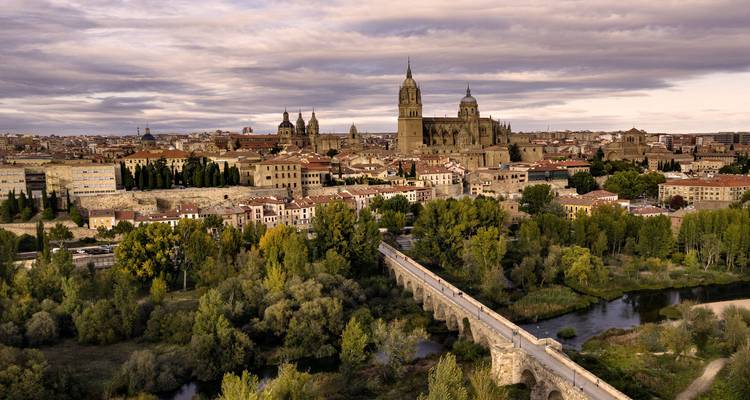A dramatic aerial view of a historic Spanish city with a stone bridge crossing a river, leafy trees in the foreground and twin–towered cathedral dominating the skyline under a moody sunset sky.