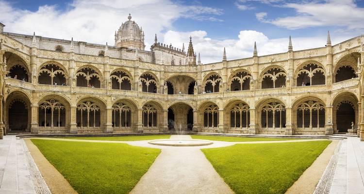 Wide-angle shot of an ornate cloister courtyard with arched stone arcades, manicured grass and a historic monastery tower rising against a bright sky.