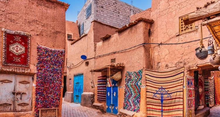 Narrow adobe alley lined with colourful Berber carpets and blue doors in a traditional Moroccan village under clear skies.