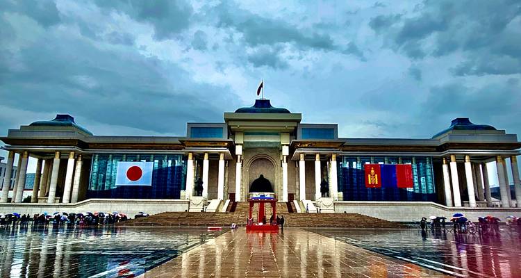 Vista frontal de un edificio grande con la bandera de Mongolia, personas con paraguas.