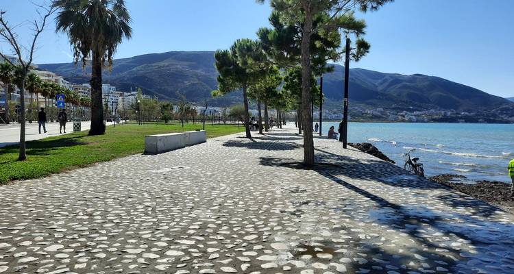 Paseo pavimentado junto al mar con árboles y montañas al fondo