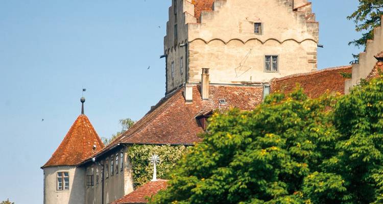 Mittelalterlicher Steinburgenturm, der sich über roten Ziegeldächern und grünen belaubten Bäumen unter einem blauen Himmel erhebt.