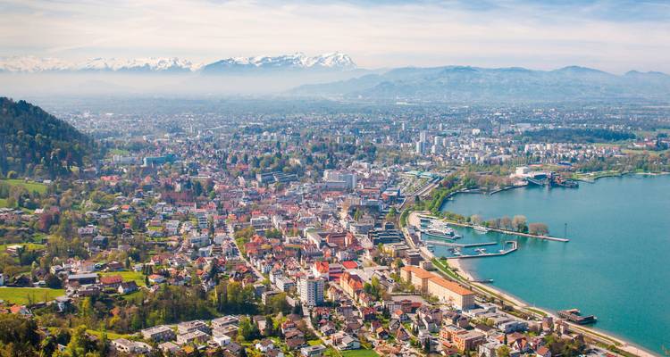 Luftaufnahme des Panoramas der Stadt Bregenz, die sich an das Ufer des Bodensees schmiegt, mit schneebedeckten Alpen in der Ferne.