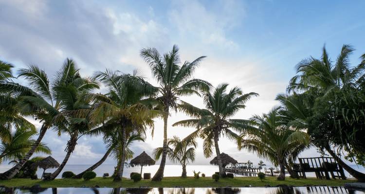 Siluetas de palmeras y cabañas contra un cielo nublado junto a una piscina.