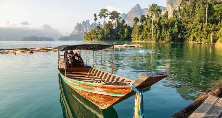 Traditional long-tail boat moored on emerald water with dramatic limestone peaks in Khao Sok National Park.
