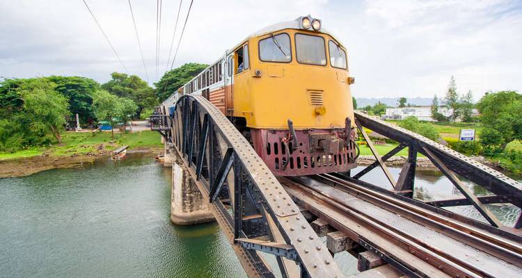 Yellow locomotive crossing a historic steel bridge over a wide river surrounded by green trees.