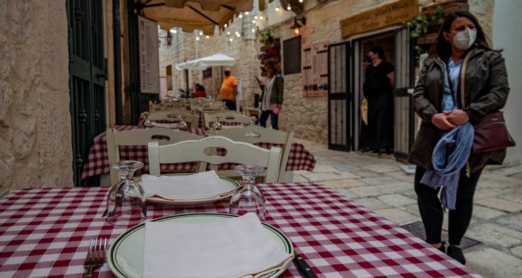 Trattoria en plein air avec des tables à carreaux rouges et blancs disposées le long d'une ruelle étroite pavée de pierre, des clients et des passants à proximité.