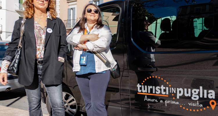 Deux femmes se tiennent à côté d'un minibus de visite touristique noir portant la marque « Turisti in Puglia » sur une place ensoleillée.