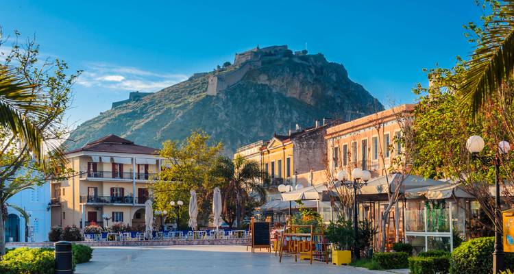 Farbenfrohe Gebäude und Café-Terrassen säumen Nafplions Hauptplatz, während die Palamidi-Festung auf einem Hügel dahinter aufragt