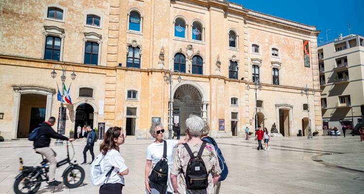 Un petit groupe se tient dans une piazza italienne ensoleillée discutant de l'histoire locale devant un bâtiment civique ocre.