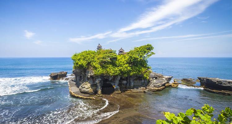 El templo marino Tanah Lot encaramado en un islote rocoso rodeado de olas oceánicas que se rompen bajo un cielo azul despejado.