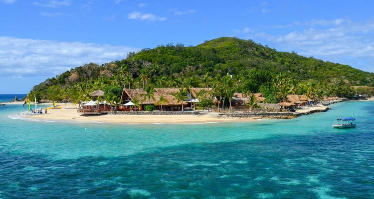 A small palm-fringed island resort is surrounded by vivid blue water and coral reefs.