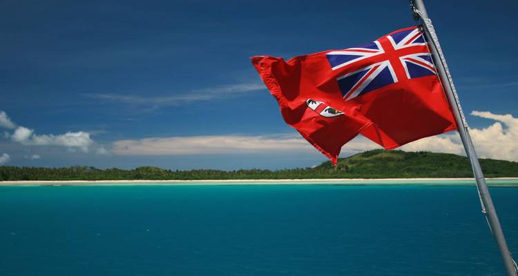 A red Fiji maritime flag flutters against a deep blue sea and distant island shoreline.