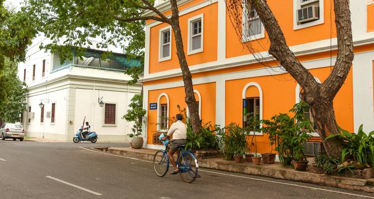 Calle colonial tranquila en Puducherry con un ciclista pasando junto a edificios patrimoniales de vibrantes colores naranja y blanco.