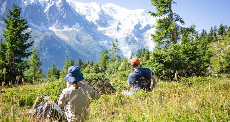 Dos excursionistas se relajan en un prado alpino lleno de flores silvestres con picos nevados a lo lejos.