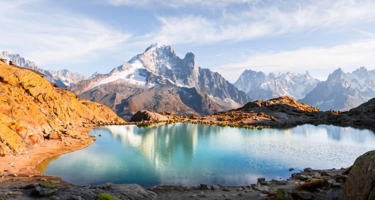 Lago de montaña cristalino que refleja picos dentados y rocas de colores otoñales.