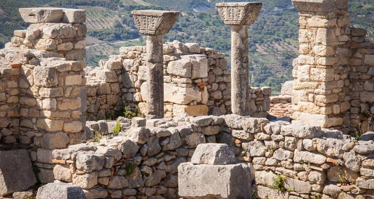 Antiguas ruinas de piedra con columnas y paisaje montañoso de fondo.