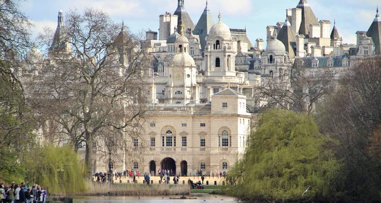 Historic Horse Guards building seen across a lake framed by leafless trees and spring foliage with visitors along the shore.