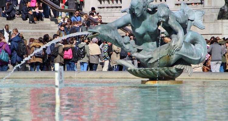 Close-up of a bronze fountain statue spraying water in Trafalgar Square with a crowd gathered on the steps behind.