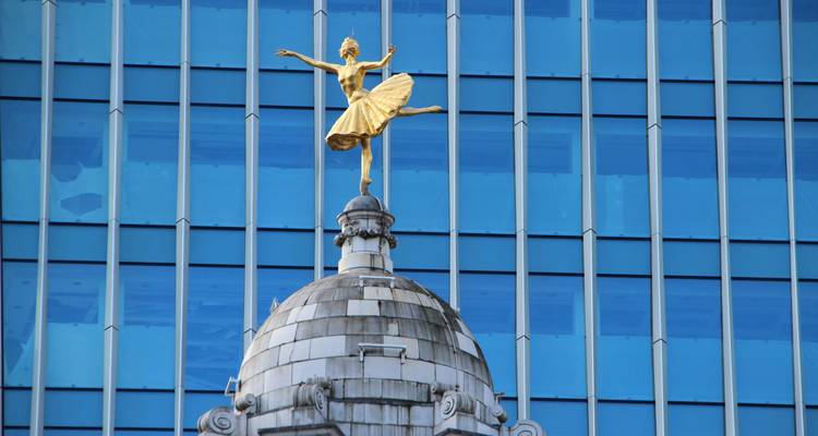 Golden ballerina statue poised atop a domed roof with a modern blue-glass skyscraper backdrop in London’s West End.
