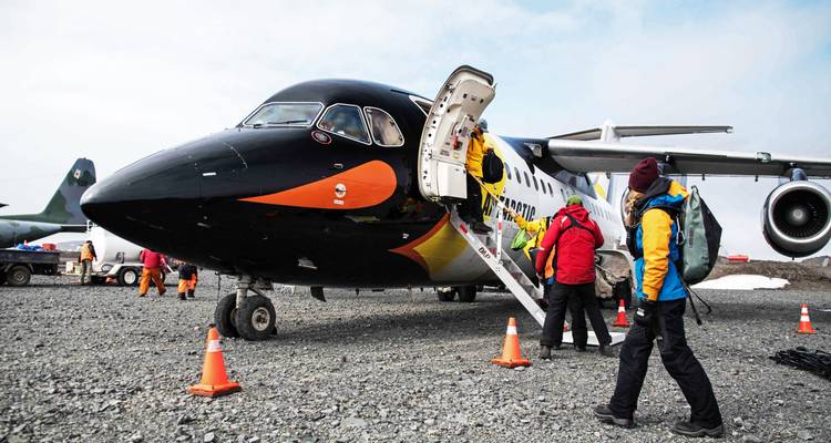Travellers descend stairway of an expedition aircraft parked on a gravel strip in polar region.