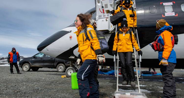 Expedition crew and passengers exit aircraft onto icy gravel runway carrying luggage and gear.