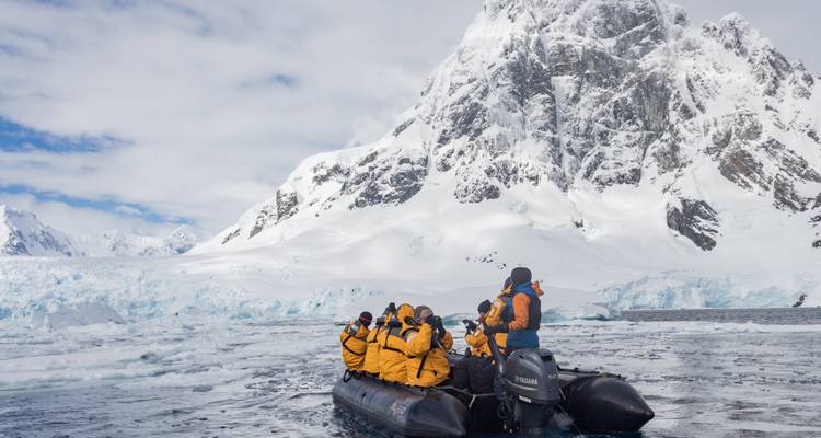 Tourists in yellow parkas ride a black inflatable Zodiac boat amid floating ice with a dramatic snow-covered mountain backdrop.