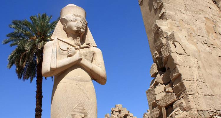 Estatua antigua de arenisca de un faraón se yergue alto junto a una palmera y muros de templo en ruinas contra un cielo azul profundo