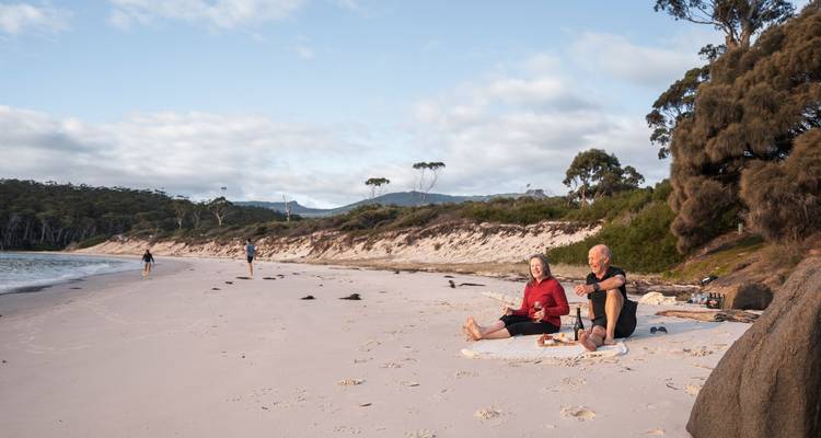 Un couple assis sur une couverture déguste du vin et des collations sur une large plage tranquille tandis que d'autres promeneurs se promènent le long du rivage.