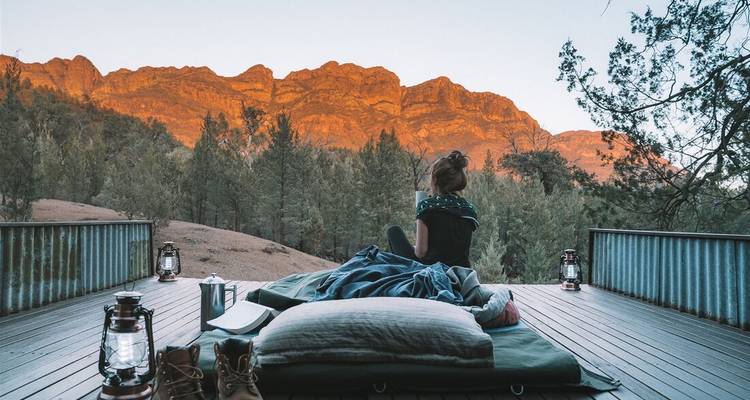 Un voyageur se détend sur un lit de plateforme extérieure à l'aube, regardant vers les chaînes de roches rouges illuminées par le soleil dans les Flinders Ranges.