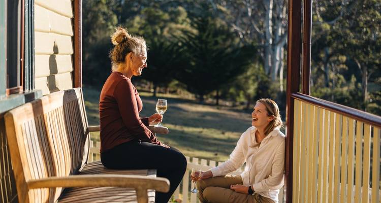 Deux femmes partageant du vin blanc sur une véranda ensoleillée surplombant un paysage rural.