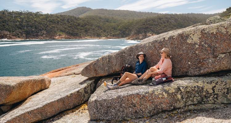 Two travellers relaxing on large coastal boulders with forested hills and surf below.