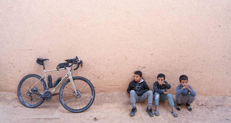Drei einheimische Jungen sitzen an einer Lehmziegelmauer neben einem geparkten Gravel-Bike.