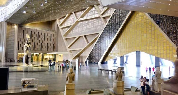 Spacious atrium of a modern museum with a towering ancient Egyptian statue, geometric ceiling panels and small groups of visitors below