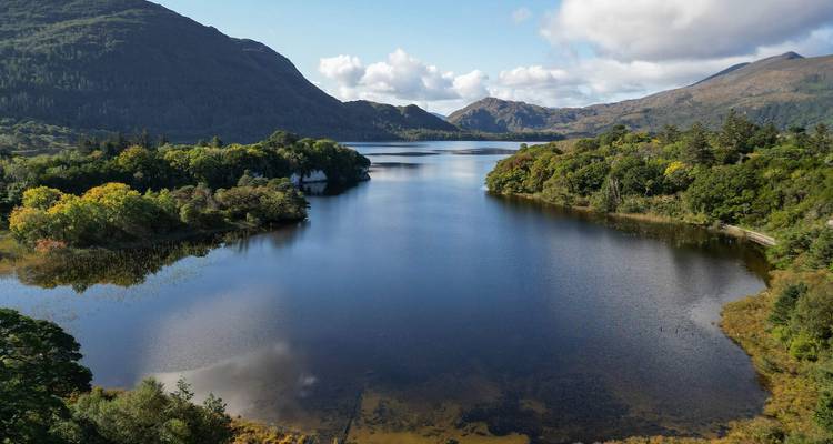 Lac serein flanqué de collines boisées et de montagnes accidentées au loin sous un ciel partiellement nuageux.