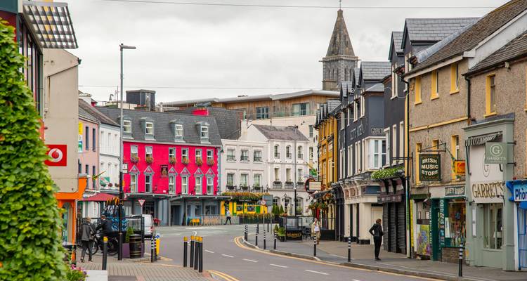 Des façades de magasins colorées bordent une tranquille rue irlandaise avec un piéton isolé et un clocher d'église au loin.