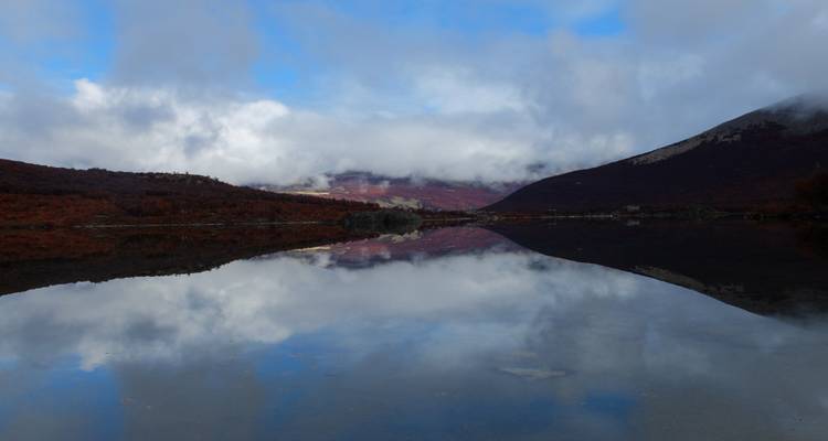 Calm reflective lake with autumnal red hills and low clouds.