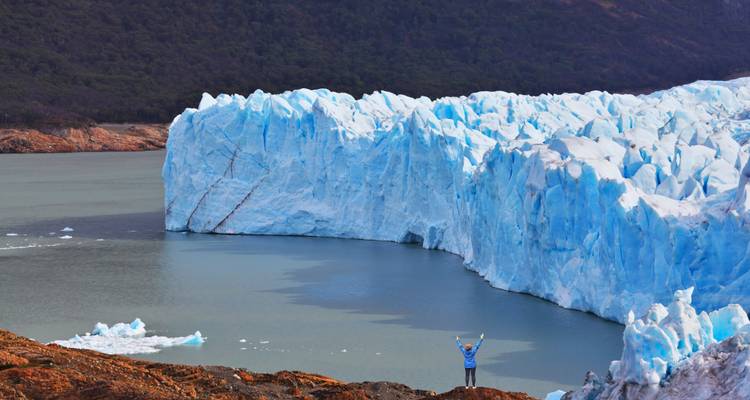 Traveller raising arms beside towering blue ice walls of a glacier.