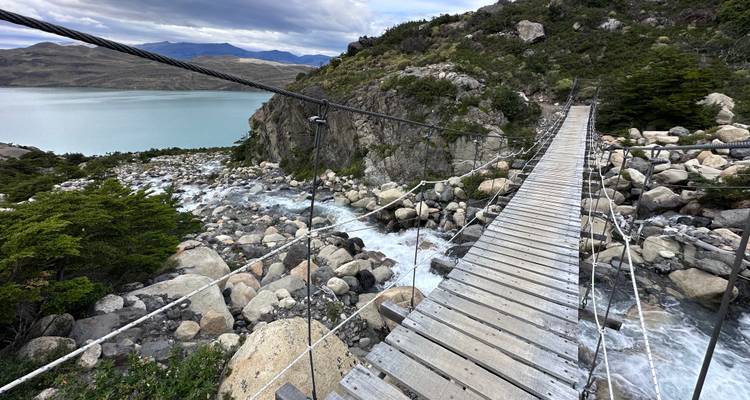 Puente colgante de suspensión cruza río impetuoso hacia lago turquesa en el paisaje de la Patagonia