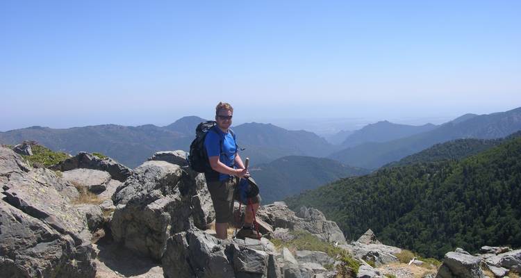 Hiker posing on rocky summit overlooking forested mountains and distant valleys.