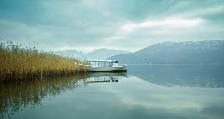 Serene lake scene with lone white boat moored beside tall reeds and misty mountains.