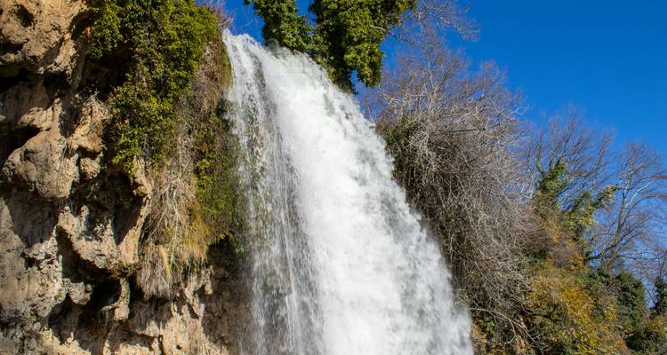 Powerful waterfall cascading over rocky cliff surrounded by greenery and blue sky.