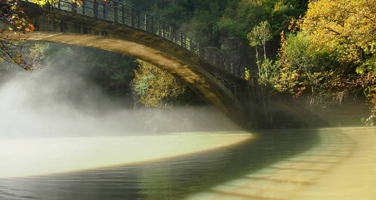 Stone arch bridge over misty river with forested backdrop in Zagori region.