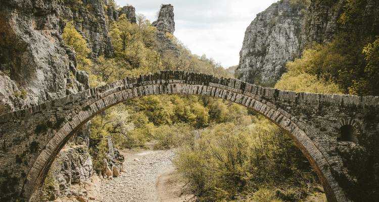 Historic stone arch bridge spanning dry riverbed in lush gorge of Zagori.