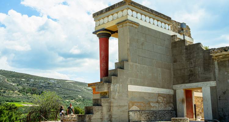 Section du palais antique de Knossos avec sa colonne rouge emblématique et des fragments de fresques sur fond de paysage rural crétois.