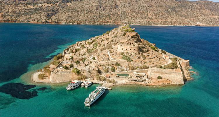 Vue aérienne par drone de la forteresse insulaire de Spinalonga avec des bateaux d'excursion amarrés à son petit quai face aux eaux bleu profond.