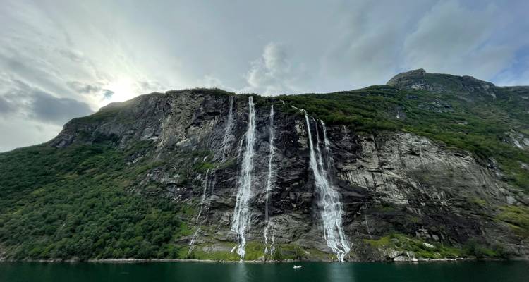 Tall, slender waterfalls cascade down a sheer cliff into emerald fjord waters beneath a cloudy sky.